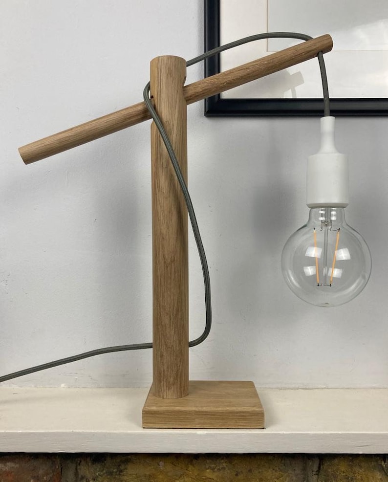 Op de afbeelding: Een houten tafellamp met een grijs snoer en een witte lamp. De lamp heeft een lange, horizontale arm die zich uitstrekt vanaf de basis.