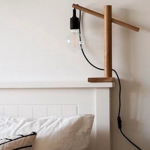 Op de afbeelding: Een houten tafellamp met een zwart snoer en een heldere gloeilamp. De lamp staat op een wit nachtkastje naast een bed met witte beddengoed.