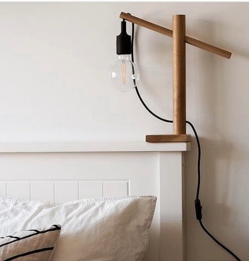 Op de afbeelding: Een houten tafellamp met een zwart snoer en een heldere gloeilamp. De lamp staat op een wit nachtkastje naast een bed met witte beddengoed.