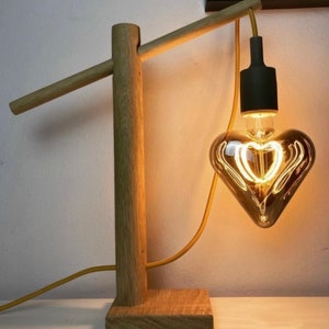 Op de afbeelding: Een houten lamp met een hartvormige gloeilamp. De lamp heeft een natuurlijke houten afwerking en een eenvoudig ontwerp. De gloeilamp is een gloeidraadlamp met een warm wit licht.