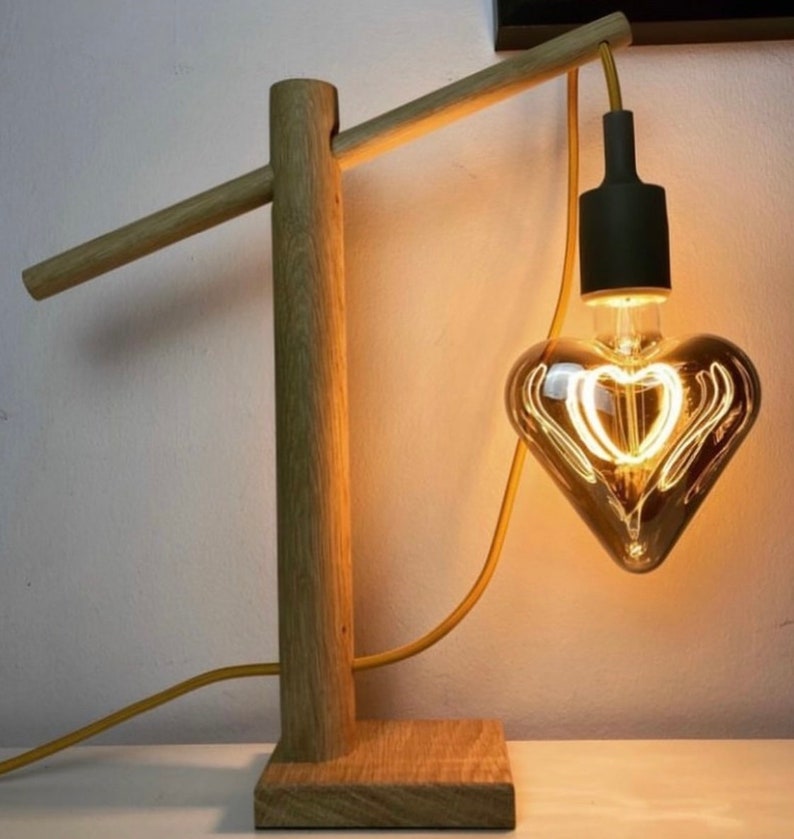 Op de afbeelding: Een houten lamp met een hartvormige gloeilamp. De lamp heeft een natuurlijke houten afwerking en een eenvoudig ontwerp. De gloeilamp is een gloeidraadlamp met een warm wit licht.