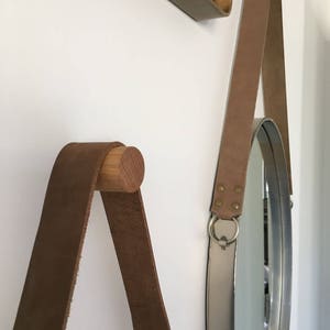 May include: Two brown leather straps with wooden dowels hang from a white wall. One strap is attached to a round mirror with a silver frame.