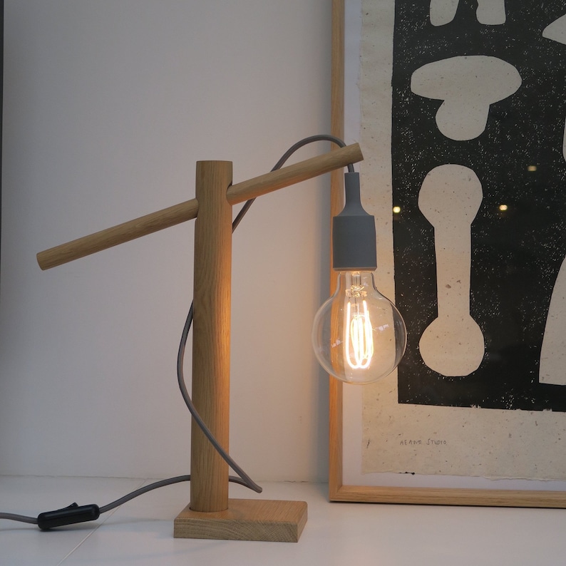 Op de afbeelding: Een houten bureaulamp met een grijs snoer en een grijze lampfitting. De lamp heeft een houten voet en een houten arm die uitsteekt om de lamp te houden. De lamp is aan en verlicht de kamer.