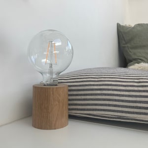 May include: A wooden table lamp with a clear glass globe light bulb. The lamp is sitting on a white surface next to a striped pillow.