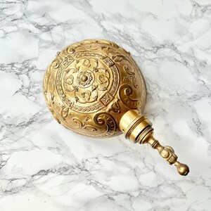 Könnte beinhalten: Eine goldfarbene Wandleuchte aus Metall mit einem verzierten, geprägten Design. Die Leuchte hat einen runden, kuppelförmigen Schirm und einen dekorativen, rollenförmigen Arm.