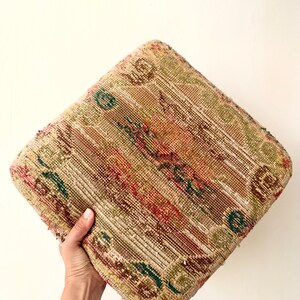 May include: A square floor cushion featuring a woven textile cover with a multi-coloured pattern in shades of tan, olive green, deep red, and dark blue. The fabric displays a textured, intricate design with visible horizontal and vertical threads.