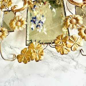 May include: Ornate gold-toned metal wall decor featuring floral and leaf motifs. The piece includes rectangular mirrored sections with hand-painted floral designs in blue, white, and green. The overall design is vintage and decorative.
