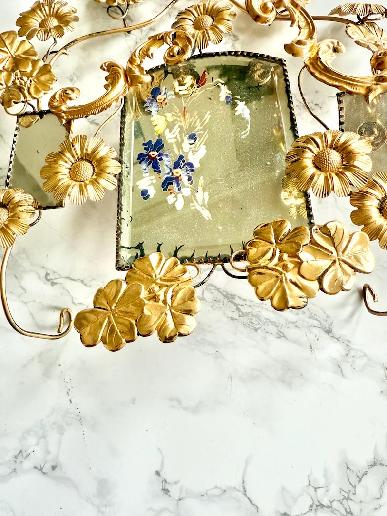 May include: Ornate gold-toned metal wall decor featuring floral and leaf motifs. The piece includes rectangular mirrored sections with hand-painted floral designs in blue, white, and green. The overall design is vintage and decorative.