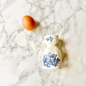 Peut inclure: Un porte-œuf en céramique blanche avec un motif floral bleu. Le porte-œuf est sur une surface en marbre blanc avec un œuf brun à côté.