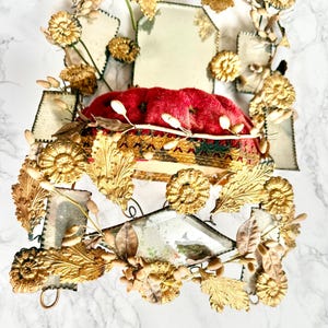 May include: An elaborate decorative item with a red velvet cushion at its centre, framed by gold-toned metal detailing. The metalwork incorporates leaf and floral designs, along with mirrored sections, giving a vintage and regal appearance.