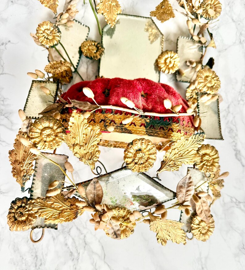 May include: An elaborate decorative item with a red velvet cushion at its centre, framed by gold-toned metal detailing. The metalwork incorporates leaf and floral designs, along with mirrored sections, giving a vintage and regal appearance.