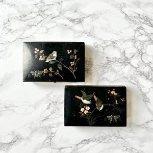 May include: Two black wooden boxes with intricate floral and bird designs. The boxes have a black background with white and gold accents. The top box features a single bird perched on a branch with flowers and berries. The bottom box features two birds on a branch with flowers and a bee. Both boxes have the letters "B" and "D" on the bottom.