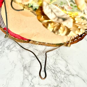 May include: A vintage, circular decorative mirror with a gold-toned metal frame and a curved metal stand. The mirror's edge is trimmed with red fabric and a patterned border. The top of the mirror is decorated with artificial flowers and leaves.
