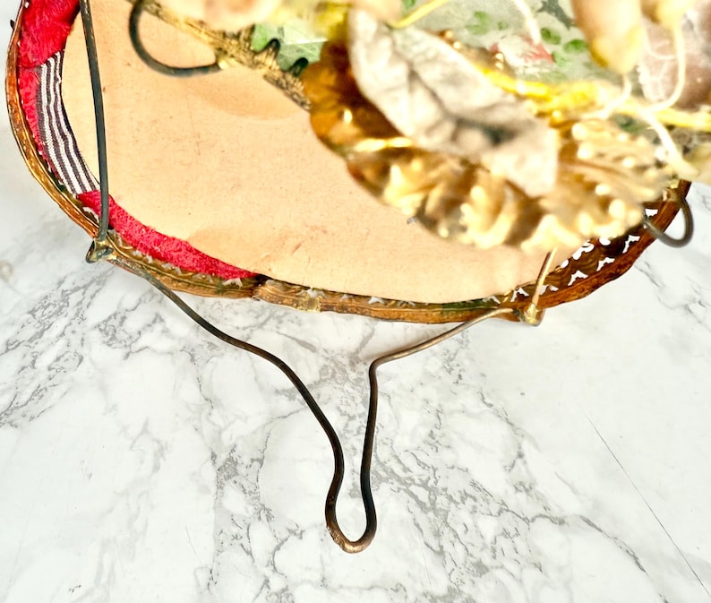 May include: A vintage, circular decorative mirror with a gold-toned metal frame and a curved metal stand. The mirror's edge is trimmed with red fabric and a patterned border. The top of the mirror is decorated with artificial flowers and leaves.