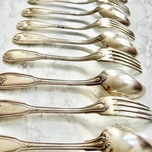 May include: A collection of silver-toned metal flatware arranged in a row, consisting of alternating forks and spoons. Each piece features a decorative shell-like motif at the base of the handle and a smooth, polished finish on the bowls and tines.