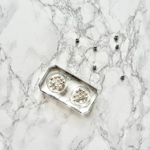 Puede incluir: Una bandeja rectangular transparente con dos compartimentos circulares, cada uno lleno de pequeñas cuentas plateadas. Varias cuentas plateadas sueltas están dispersas alrededor de la bandeja sobre una superficie de mármol blanco y gris. La bandeja tiene un diseño geométrico.