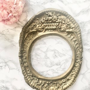 May include: An oval picture frame with ornate floral details, finished in a gold tone. The frame is displayed on a white marble surface.