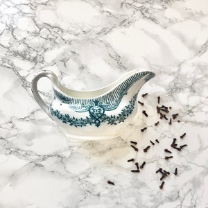 Könnte beinhalten: Eine weiße Porzellan-Soßenschüssel mit grünem Blumendesign und einem Henkel. Die Schüssel steht auf einer weißen Marmorfläche mit einer Streuung von braunen Nelken.