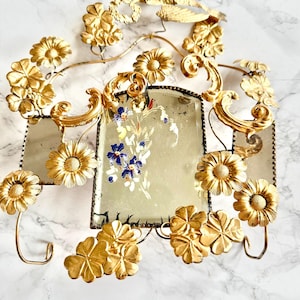 May include: Ornate gold-colored wall decor featuring floral and scroll designs. The piece includes two rectangular mirrors with a floral painting in the center. Decorative flowers and leaves are attached to the frame.