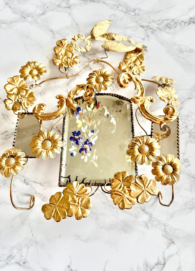 May include: Ornate gold-colored wall decor featuring floral and scroll designs. The piece includes two rectangular mirrors with a floral painting in the center. Decorative flowers and leaves are attached to the frame.