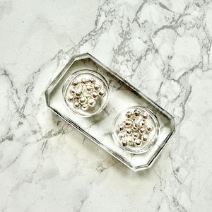 Puede incluir: Bandeja rectangular transparente con dos pozos circulares, cada uno lleno de pequeñas esferas plateadas brillantes. La bandeja tiene un borde facetado. El fondo es un patrón de mármol blanco y gris.