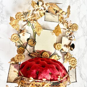 May include: An elaborate, decorative object with a plush, red cushion base and gold-coloured, detailed trim. It is decorated with gold-coloured floral and leaf accents, and a small bird figure. The background has a white marble pattern.