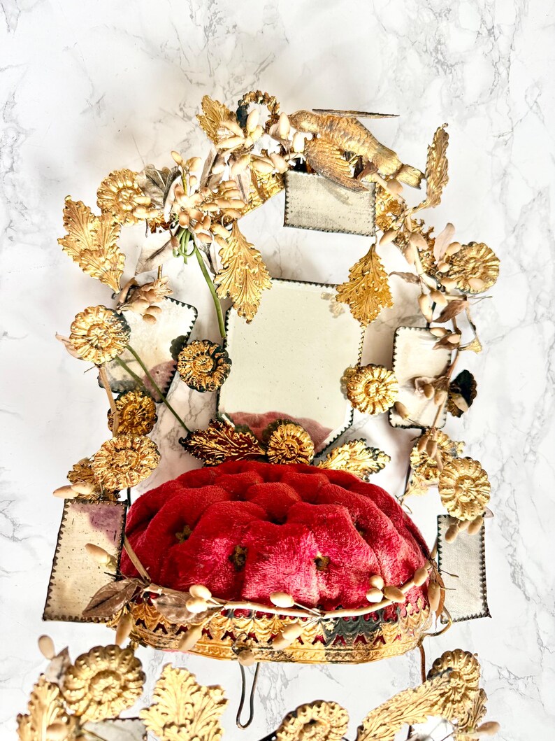 May include: An elaborate, decorative object with a plush, red cushion base and gold-coloured, detailed trim. It is decorated with gold-coloured floral and leaf accents, and a small bird figure. The background has a white marble pattern.