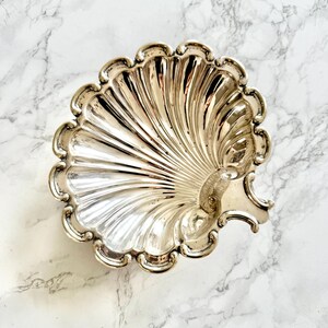 Peut inclure: Un plat argenté en forme de coquillage avec un bord festonné. Le plat a un intérieur nervuré et un petit détail en forme de poignée sur un côté. Le plat est présenté sur une surface en marbre blanc et gris.