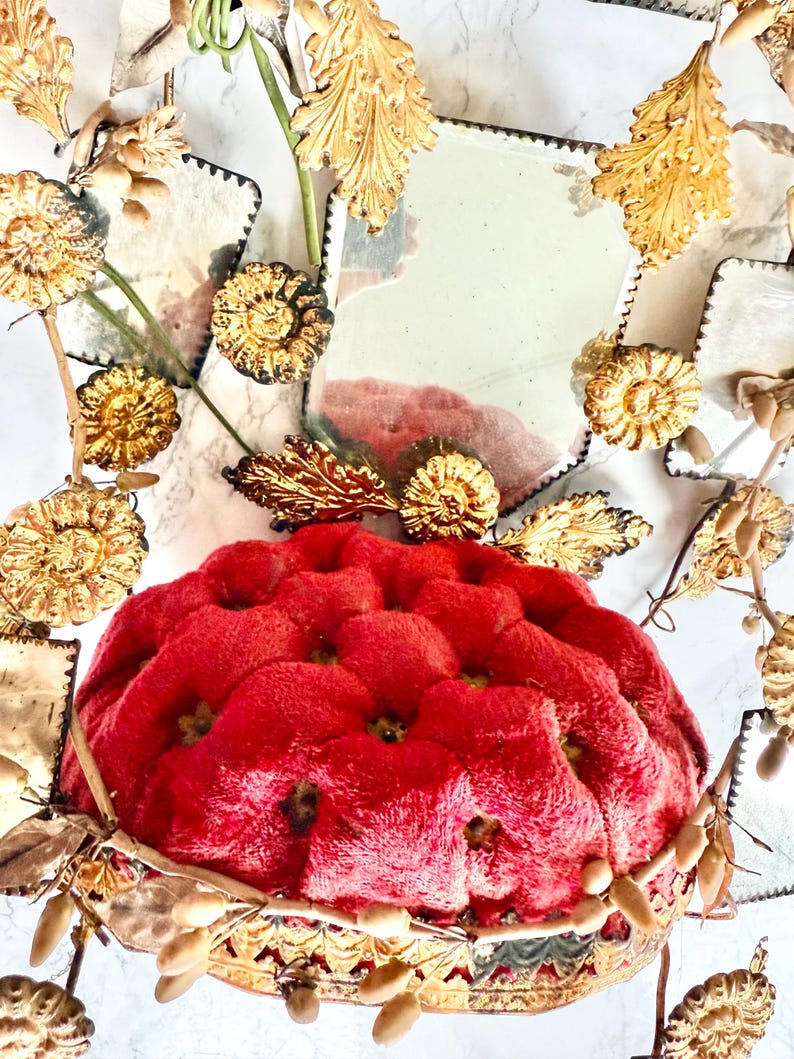 May include: A red, tufted cushion with a decorative gold-coloured base. The cushion is surrounded by gold-coloured floral and leaf accents, and small, square mirrors. The overall design is ornate and vintage-inspired.