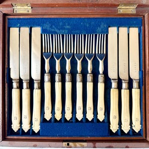 Peut inclure: Un ensemble de couverts anciens dans un coffret en bois. L'ensemble comprend des couteaux et des fourchettes avec des manches de couleur ivoire et des lames argentées. L'étui est doublé de velours bleu, les couverts sont rangés à l'intérieur.