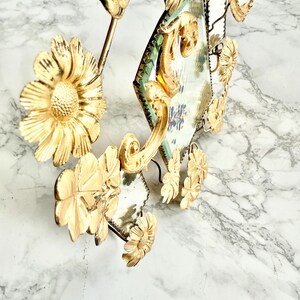 May include: Ornate gold-colored wall decor featuring floral and mirror elements. The design includes stylized flowers and diamond-shaped mirrors with a green border. The piece has a vintage aesthetic and is set against a marble background.