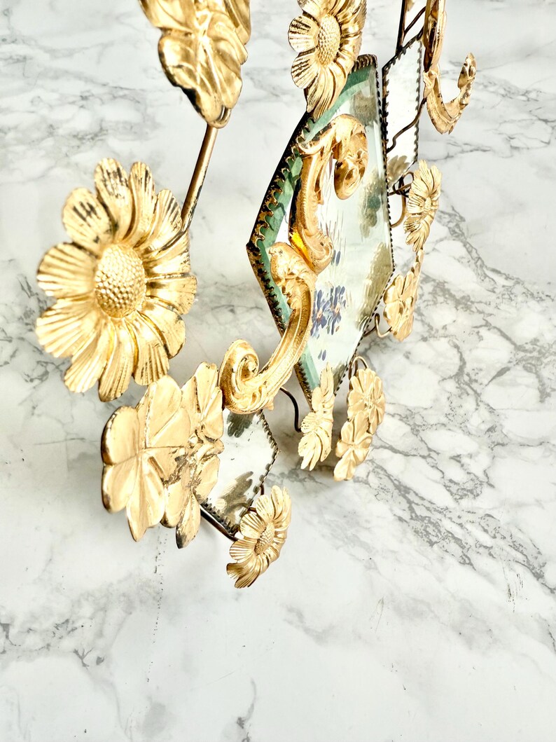 May include: Ornate gold-colored wall decor featuring floral and mirror elements. The design includes stylized flowers and diamond-shaped mirrors with a green border. The piece has a vintage aesthetic and is set against a marble background.