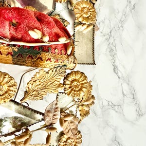 May include: An ornate gold crown with a red velvet cushion. The crown is decorated with gold floral and leaf details. The background is a white marble surface.