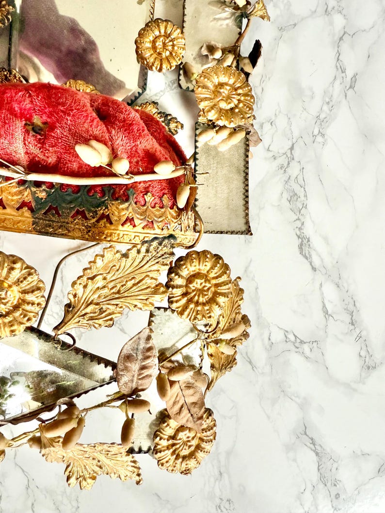 May include: An ornate gold crown with a red velvet cushion. The crown is decorated with gold floral and leaf details. The background is a white marble surface.