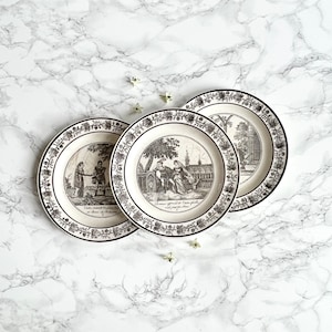 May include: Three white ceramic plates with black and white illustrations of people in a garden setting. The plates have a decorative black border with floral designs.