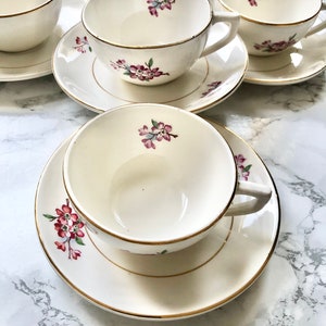 May include: Set of four white teacups and saucers with a delicate floral design in shades of pink and green. The cups and saucers have a gold trim.