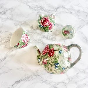 Peut inclure: Un service à thé en céramique comprenant une théière, un pot à lait, un couvercle de sucrier et une tasse. Chaque pièce est ornée d'un motif de roses roses, de feuilles vertes et de petites fleurs bleues sur un fond crème à texture pointillée.