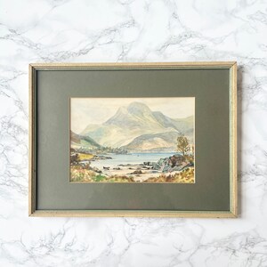May include: A framed watercolour painting depicting a scenic landscape with a lake, mountains, and trees. The painting is in a green frame with a light brown mount.