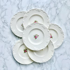 Peut inclure: Ensemble de six assiettes plates en céramique blanche avec un motif floral en relief et une seule rose rose au centre de chaque assiette.
