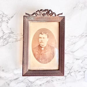 Peut inclure: Un cadre photo rectangulaire en métal avec une finition bronze foncé et une crête ornementale en forme de ruban sur le dessus. Le cadre contient un portrait ovale sépia d'une personne avec une moustache portant une veste de costume formelle.