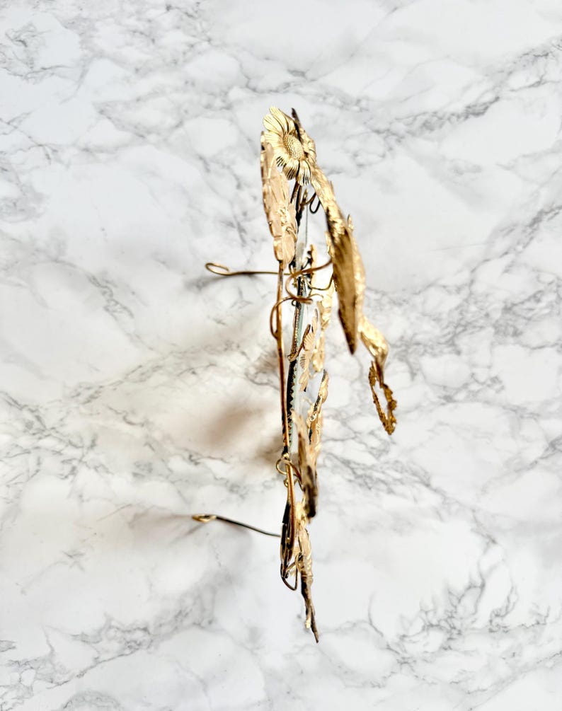 May include: A gold-colored metal floral headpiece with a sunflower and leaf design. The headpiece has a wire frame and is displayed against a marble background.