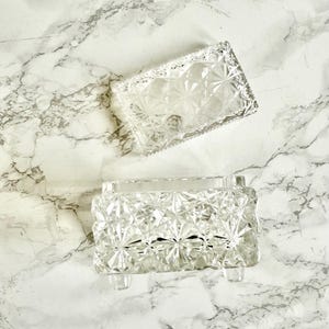 May include: Two clear, rectangular glass dishes with a diamond pattern. One dish is open, and the other is a lid. The dishes are on a white and gray marble surface. The dishes have a vintage aesthetic.