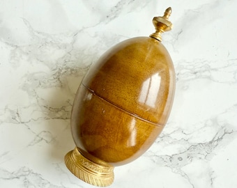 May include: An egg-shaped wooden container with a polished brown finish, featuring a metallic gold-toned base with a textured pattern and a matching gold-toned finial topper that sits atop the rounded wooden lid.