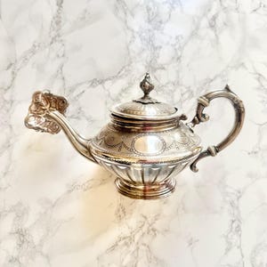 Peut inclure: Une théière en argent avec des détails ornés, notamment un bec verseur, une anse et un bouton de couvercle décoratifs. La théière présente des motifs gravés et un corps arrondi, reposant sur une base circulaire. Le fond est une surface en marbre.