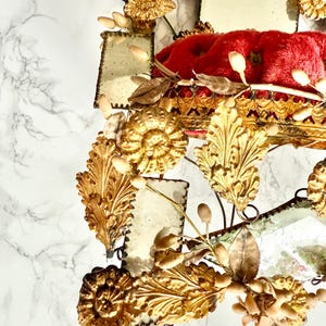 May include: An antique mirror with a red velvet cushion and gold-toned embellishments. The mirror features intricate floral and leaf designs, with small rectangular mirror panels. The frame is detailed with gold accents and a vintage aesthetic.