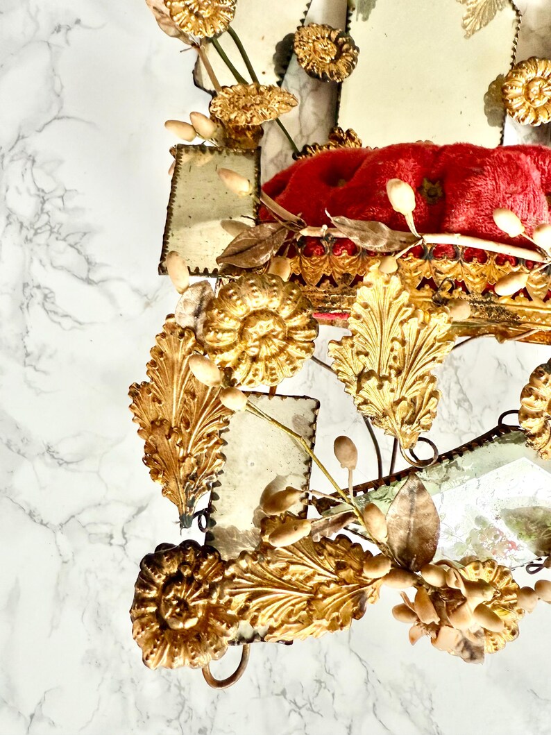 May include: An antique mirror with a red velvet cushion and gold-toned embellishments. The mirror features intricate floral and leaf designs, with small rectangular mirror panels. The frame is detailed with gold accents and a vintage aesthetic.