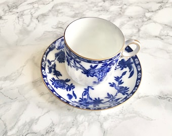 May include: A white teacup and saucer with a blue and gold floral pattern. The cup has a gold rim and the saucer has a gold band around the edge.