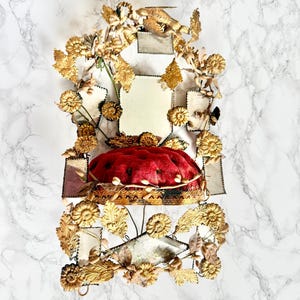 Peut inclure: Une pièce décorative dorée ornée d'un coussin rouge central. Le coussin est entouré d'éléments floraux et de feuilles dorées, de petits miroirs et d'éléments rectangulaires. Le design est symétrique et détaillé, avec une esthétique vintage.