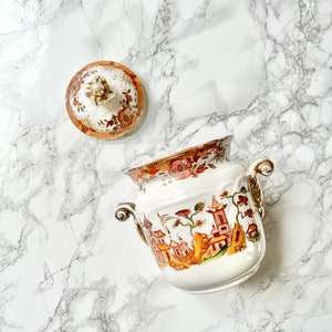 Può includere: Una zuccheriera in ceramica bianca con coperchio, con un design dipinto a mano di un paesaggio cinese in tonalità di rosso, arancione e verde. La ciotola ha due manici dorati e un bordo dorato attorno al bordo.