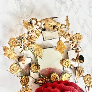 May include: A decorative mirror with a gold-coloured frame and a red, tufted cushion base. The frame is adorned with detailed floral and bird-like elements. The mirror's surface is divided into several small, square sections.